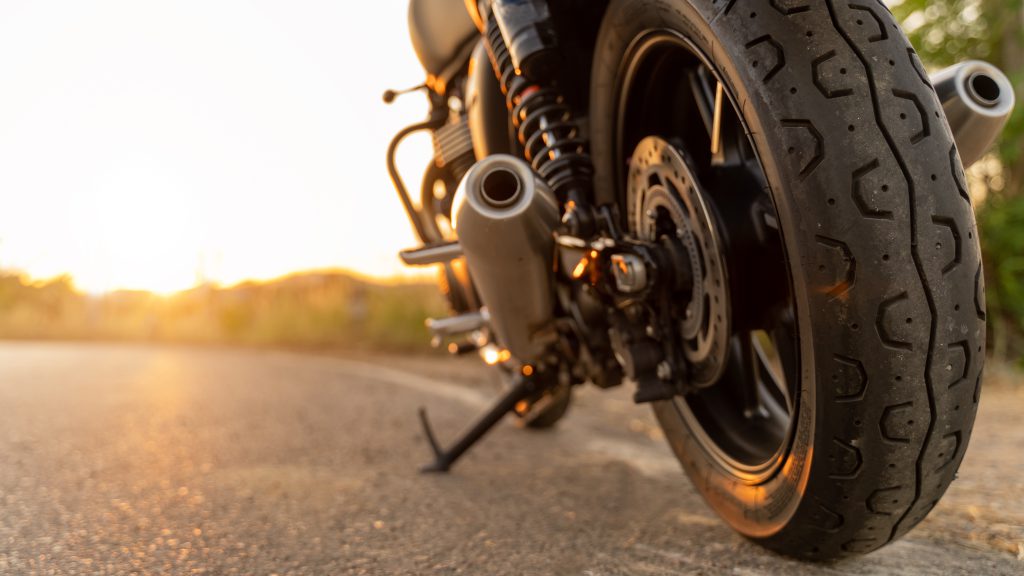 Rear end of a motorcycle facing a sunset whilst parked on the side of a road.